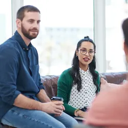 A couple sitting comfortably on a couch during a therapy session, the woman wearing a polka dot shirt and green cardigan while the man holds a coffee cup, both engaged in conversation with their therapist