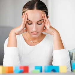 A stressed woman is holding the sides of her head in her hands while staring at blocks on a table