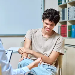 A smiling teenage boy is sitting across from a mental health counselor while listening to her speak